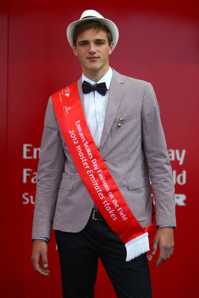 Person wearing a patterned suit, bow tie, and fedora, with a "2019 Epsom Derby - Stewards' Emirates Melbourne Cup" sash