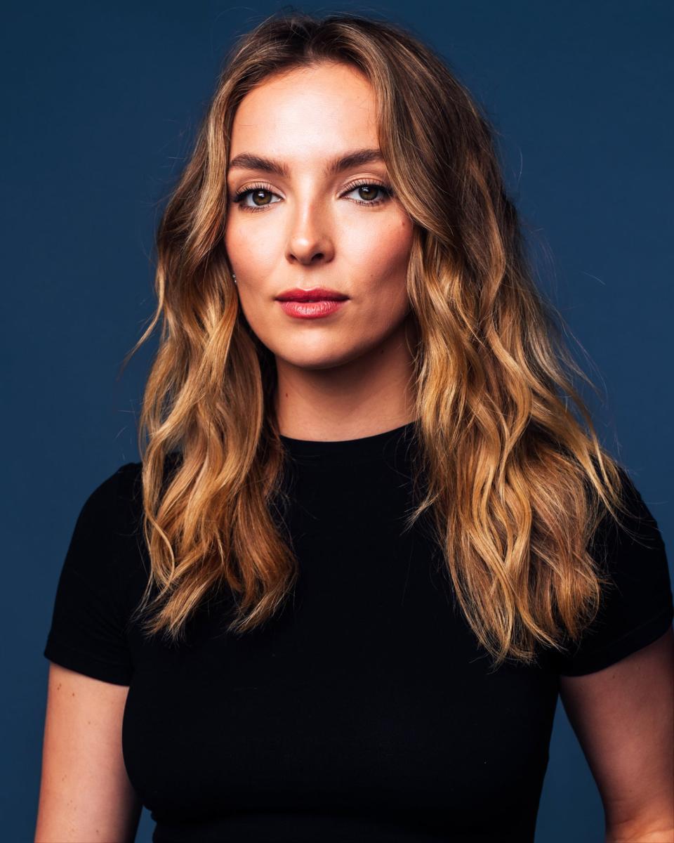 Jodie Comer with wavy hair in a shoulder-length hairstyle is wearing a black top and looking directly at the camera