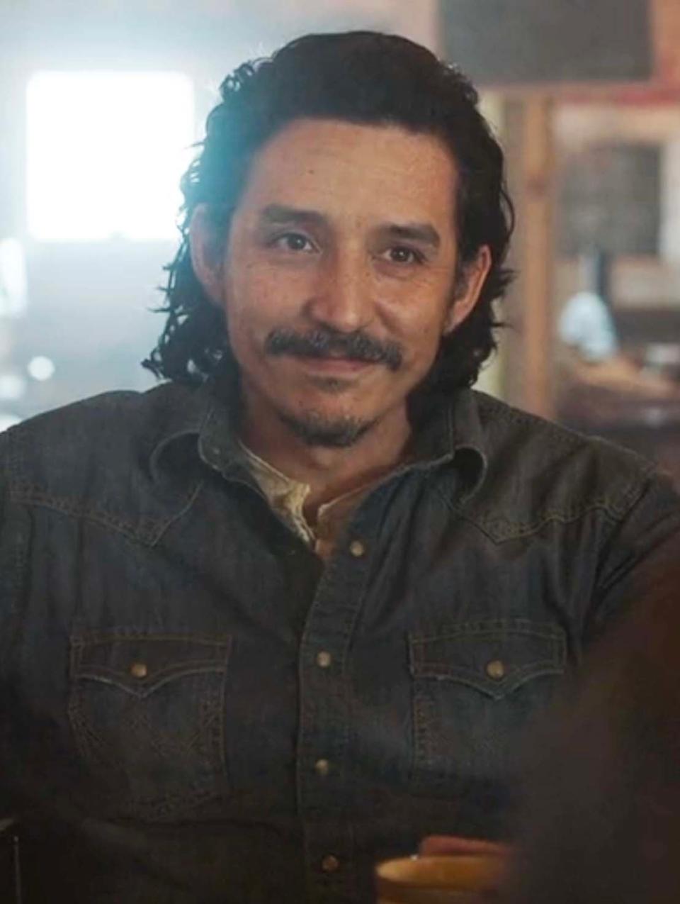 Tommy in The Last of Us Season 1 in a denim shirt sitting, slightly smiling. Appears in a casual indoor setting