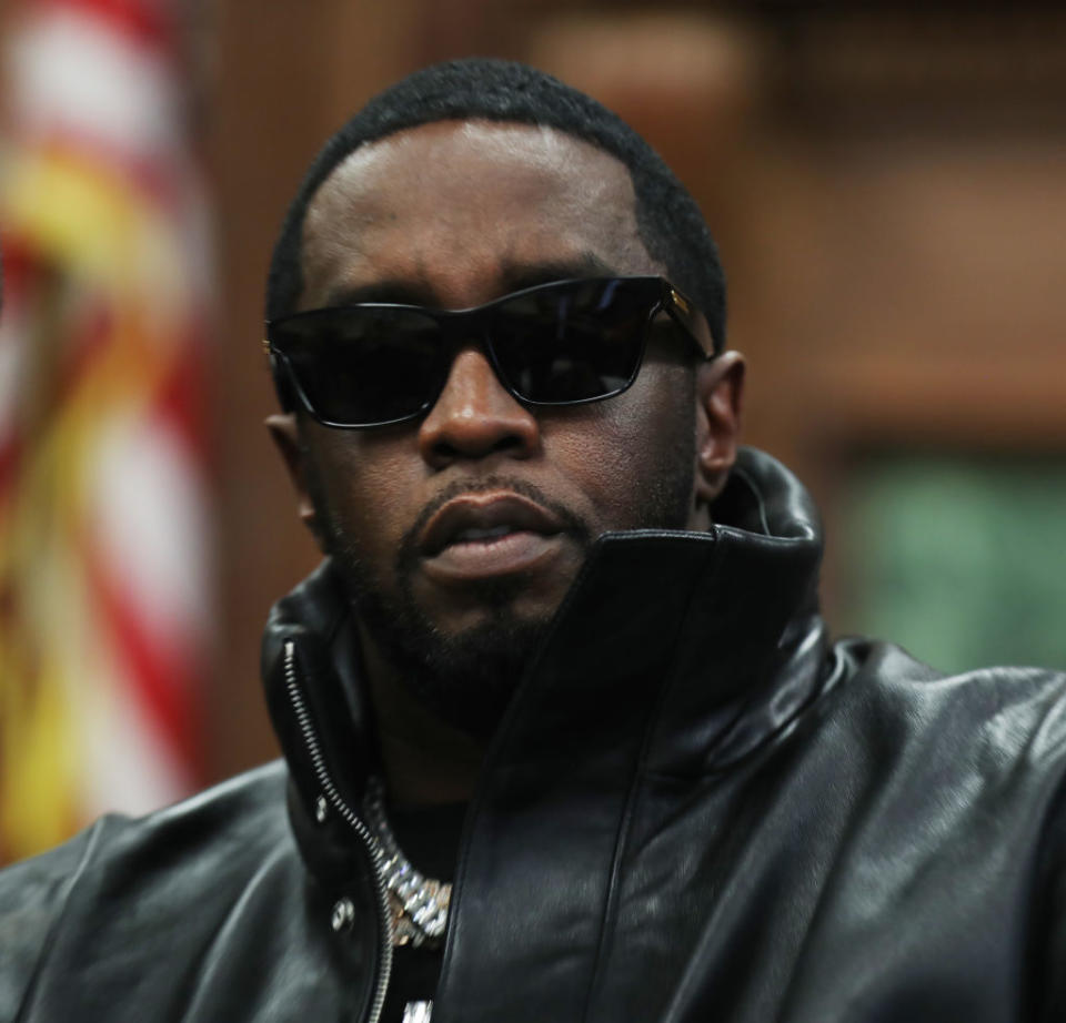 Sean Combs, wearing dark sunglasses and a leather jacket, stands in front of an American flag and appears serious