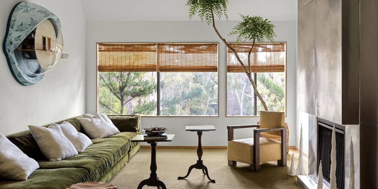 living room with high ceilings and a long sofa covered in a plant green velvet against the left wall and several small tripod tables scattered at center and opposite the sofa is a built in fireplace clad in metal and windows on the back wall with bamboo shades rolled up and a tall plant or tree peeking out from the corner of the room