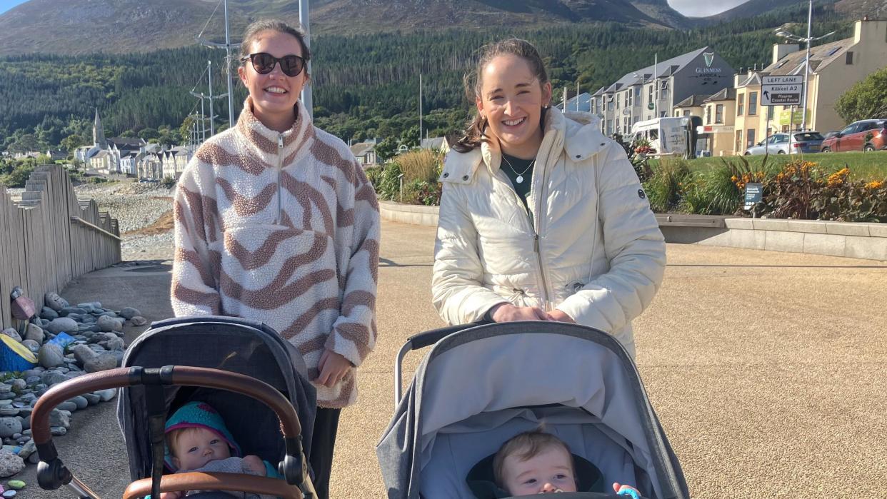 Aisling Doran and Maria are smiling at the camera with their babies in prams.

Behind them is the Mourne Mountains. 

The sun is shining and there are trees behind them and houses to the right and a pebbly beach to the left. 

Aisling is wearing a fleece with white and maroon patterns on it and Marie is wearing a white puffet jacket. 