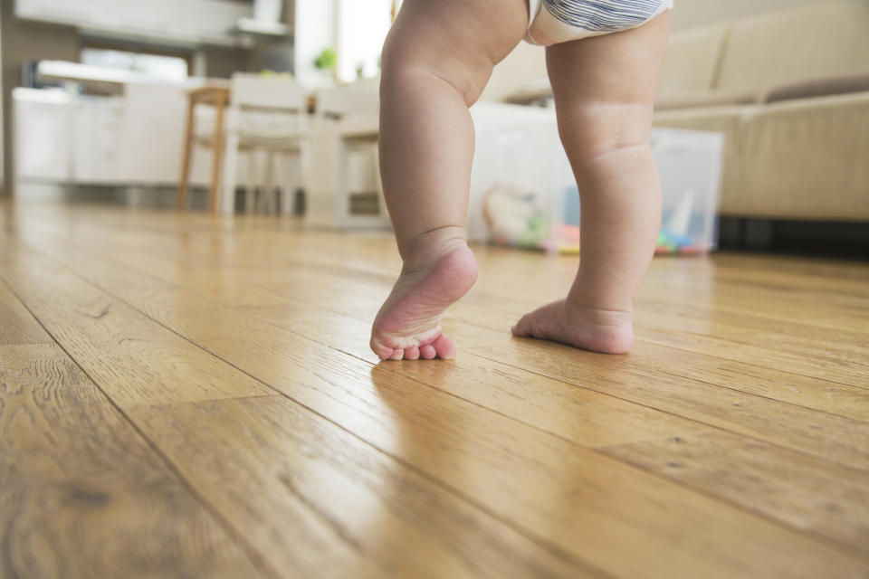 A toddler's legs are visible as they learn to walk in a home environment with wooden floors