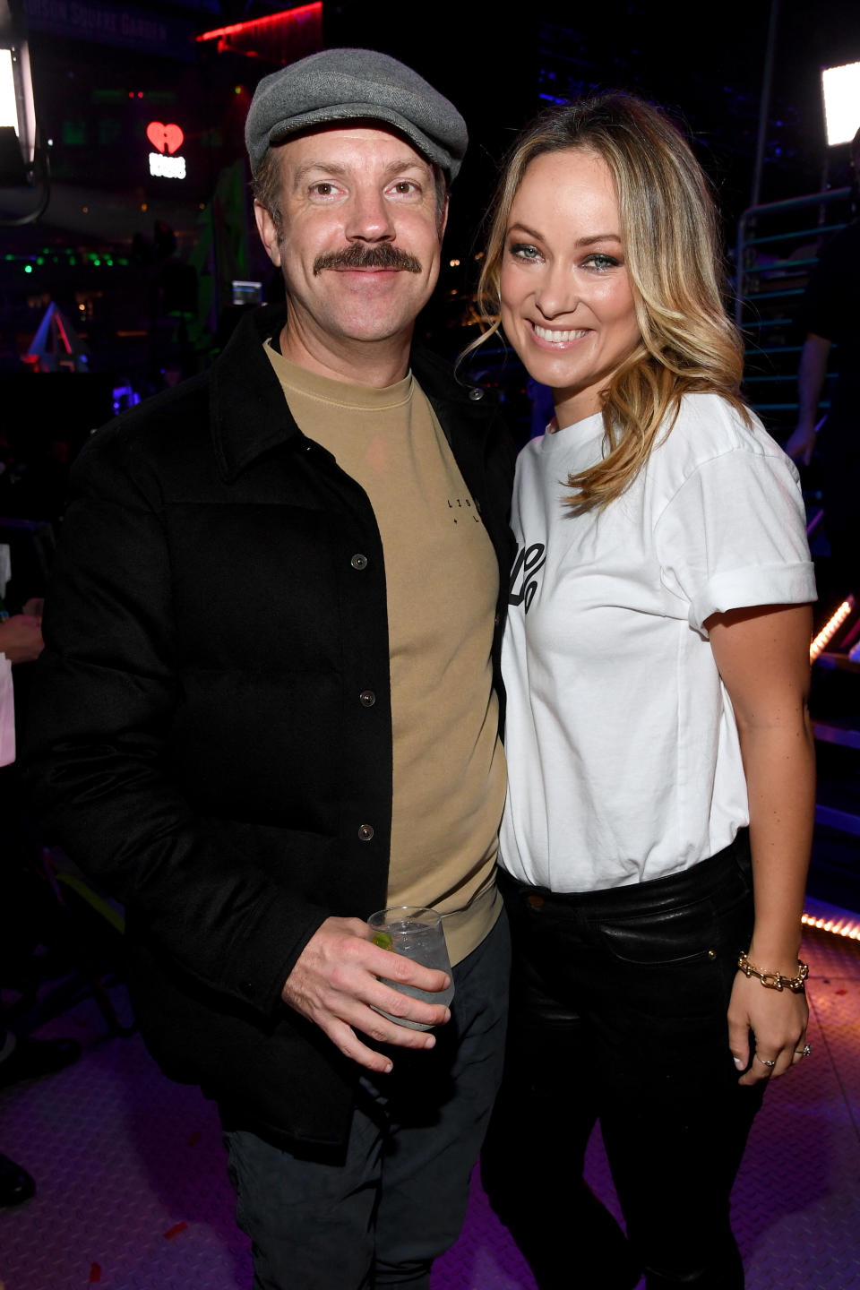Two people posing for a photo, man in a cap and jacket, woman in a white tee and black pants