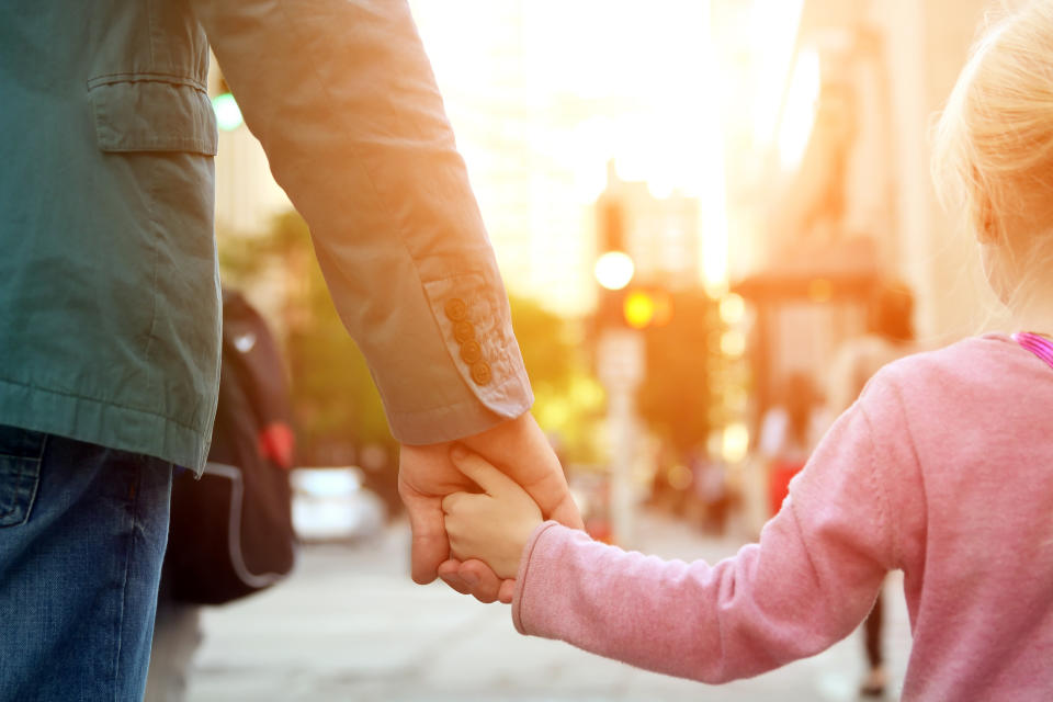 A man and a young girl holding hands while walking down a city street. Their faces are not visible. The girl is wearing a long-sleeve shirt