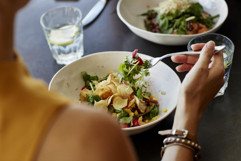 Person eating a salad with chips, seen over the shoulder, at a table with another meal and water glasses