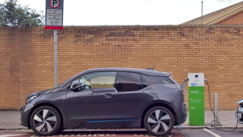 A hatchback electric vehicle charges at a public charger adjacent to a low brick building.