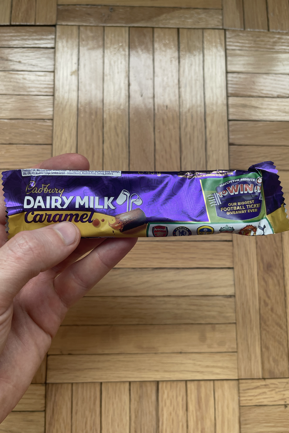 Hand holding a Cadbury Dairy Milk Caramel chocolate bar on a wooden floor