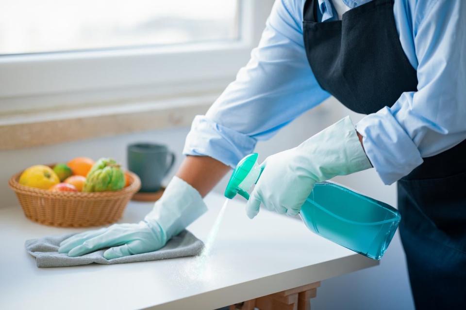 A person in a blue outfit and cleaning gloves sprays a solution on a countertop and wipes it down. 