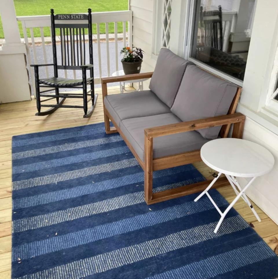 The wooden loveseat with cushions on front porch with a rocking chair, and white side table, all on a striped rug