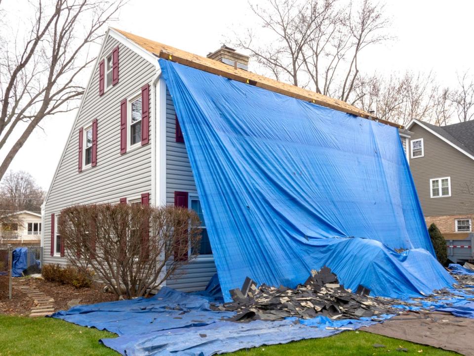 grey colonial home covered in large blue tarp with roof tiles on lawn while roof is being replaced