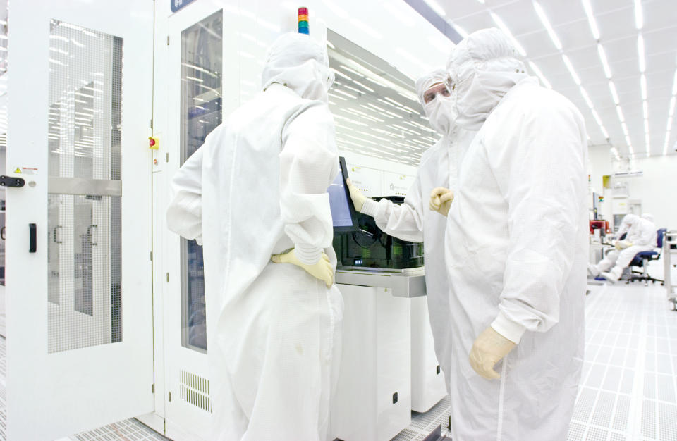 Three workers in head-to-toe white lab suits confer in a laboratory.