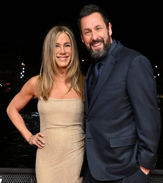 Jennifer Aniston in a sequined gold dress puts her right hand on her hip and smiles for a photo with Adam Sandler wearing a navy suit and tie