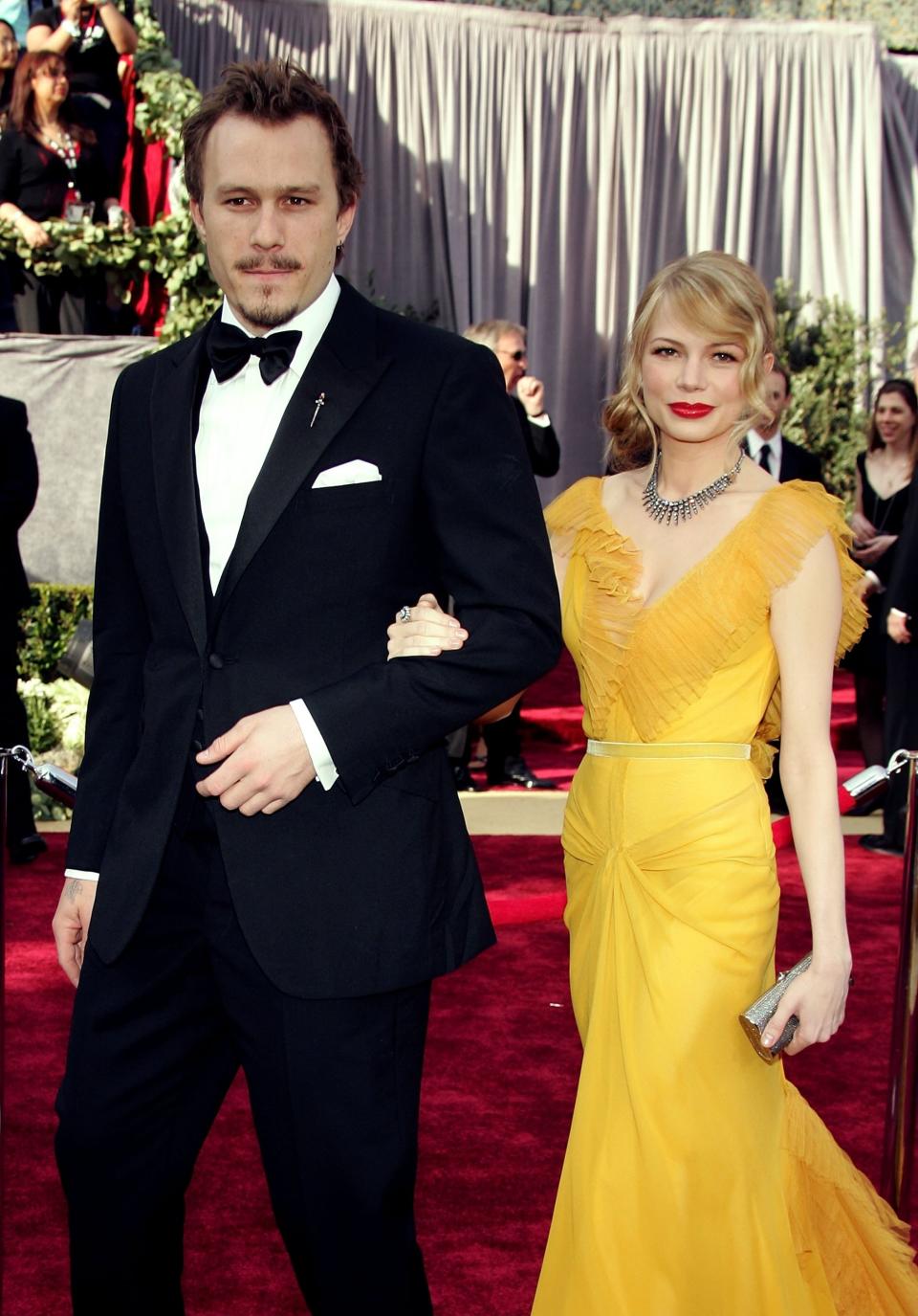 Heath Ledger and Michelle Williams on the red carpet. Heath is in a black tuxedo, and Michelle is in a yellow gown with a deep neckline and ruffled details