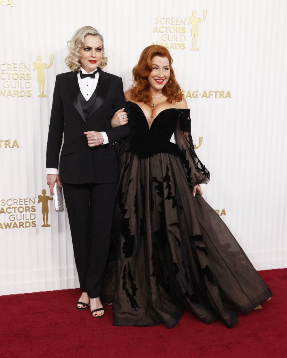 A blond woman in a suit and a red-haired woman in a gown linking arms and laughing on a red carpet.