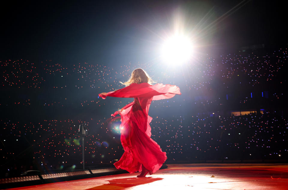 Taylor Swift in flowing red dress on stage with bright lights and audience in background
