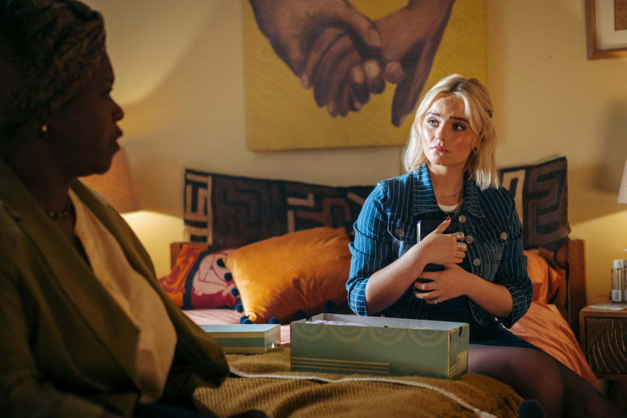  Doctor Who: Ruby Sunday (Millie Gibson) sits on her mum's bed, with an open shoebox in front of her. Ruby is clutching something from inside the box to her chest, as her mother Carla (Michelle Greenidge) - visible in the foreground but out of focus - is talking to her. 