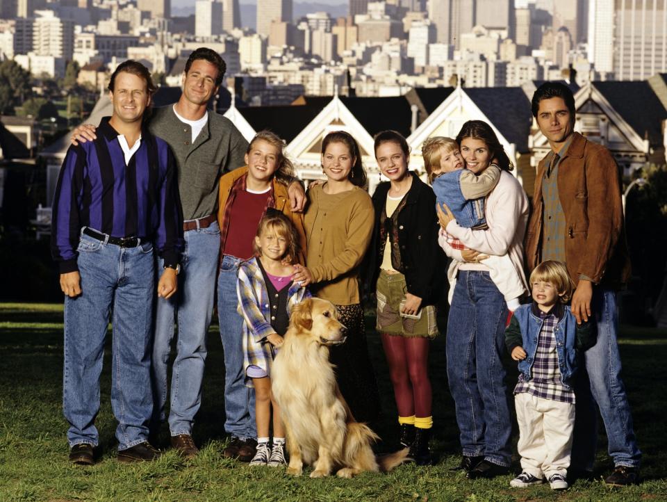 The image shows the cast of "Full House" posing outdoors with the San Francisco skyline in the background. Members include Bob Saget, Dave Coulier, John Stamos, and others