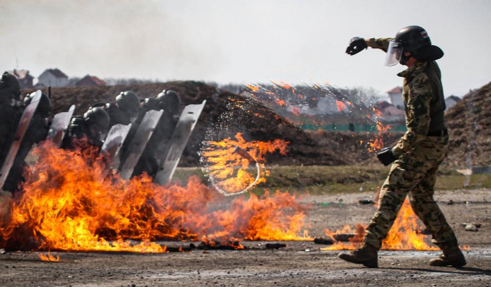 A photo of a soldier in a face shield and helmet throwing a spinning Molotov cocktail at a group of soldiers.