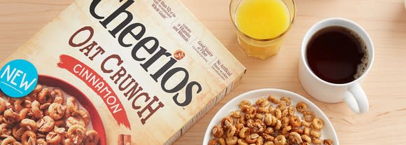 Box of Cheerios Oat Crunch Cinnamon cereal, with filled bowl, orange juice cup, and coffee cup on wooden table