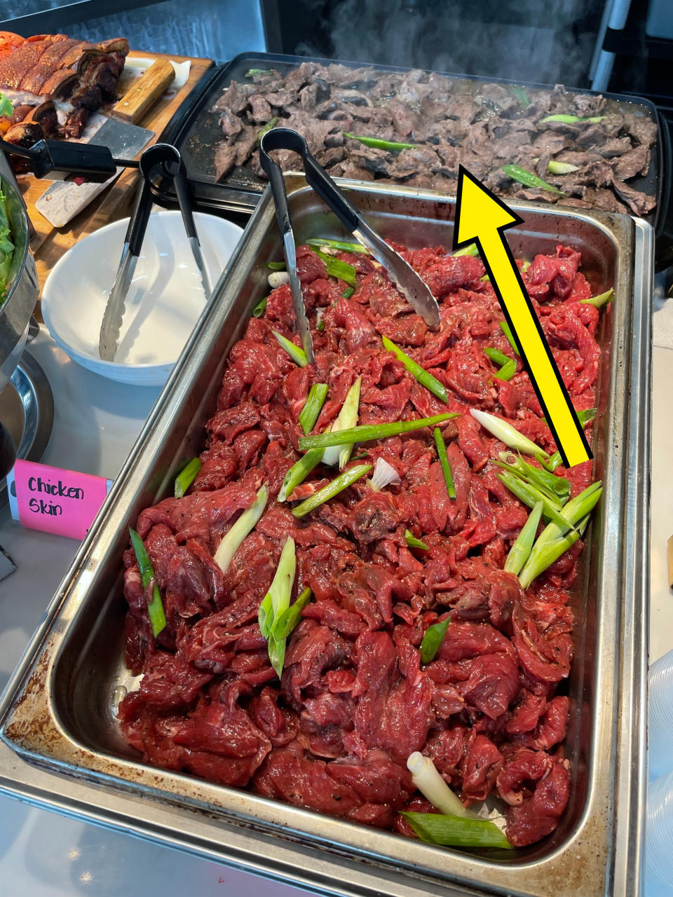 A buffet of sliced raw beef with green onions and cooked beef in separate trays is displayed. There is also a container labeled "chicken skin."