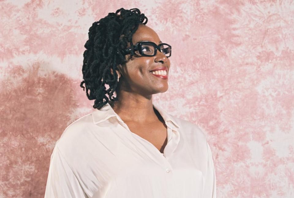 portrait of Dawn Barker-Pierre, middle aged black woman with black glasses and black hair wearing white blouse in front of pink pattern wall