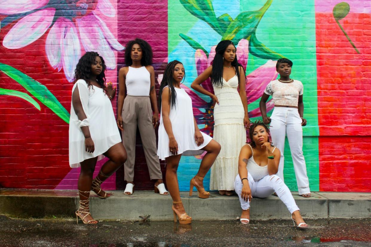 six black women in beige and white in front of a colorful brick wall