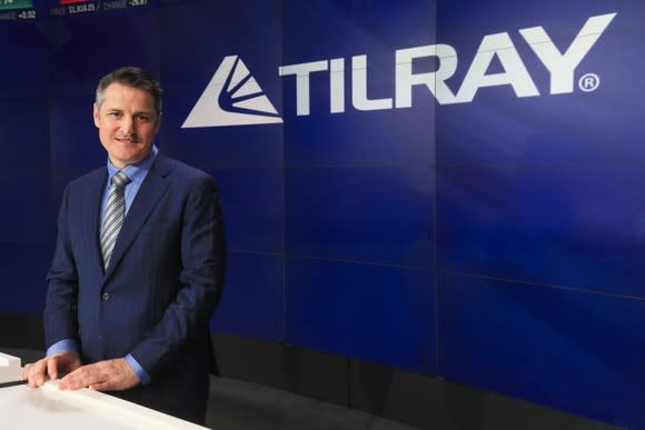 Person wearing suit standing at a desk in front of blue screen with Tilray logo on it.