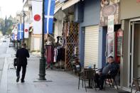 A local resident sits at a cafe as a Japanese official walks in the town of Olympia, southern Greece, Wednesday, March 11, 2020. Greek Olympic officials in ancient Olympia, birthplace of the ancient Olympics are holding a pared-down flame-lighting ceremony for the Tokyo Games due to concerns over the spread of the coronavirus. Both Wednesday's dress rehearsal and Thursday's lighting ceremony will be closed to the public, while organizers have slashed the number of officials from the International Olympic Committee and the Tokyo Organizing Committee, as well as journalists at the flame-lighting. (AP Photo/Thanassis Stavrakis)