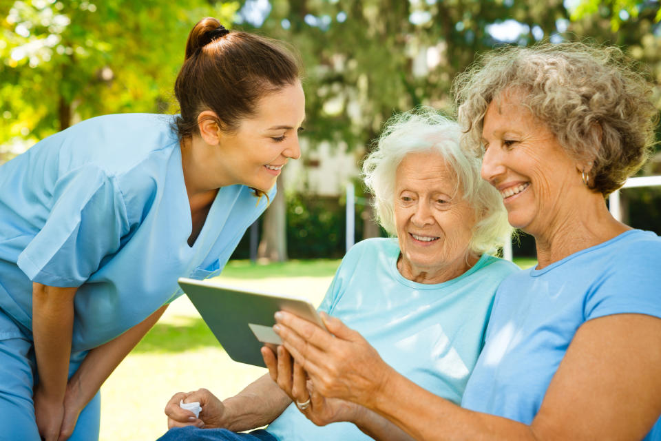 Smiling nurse looking at senior women using digital tablet. Healthcare worker is with retired females at nursing home. They are at back yard.