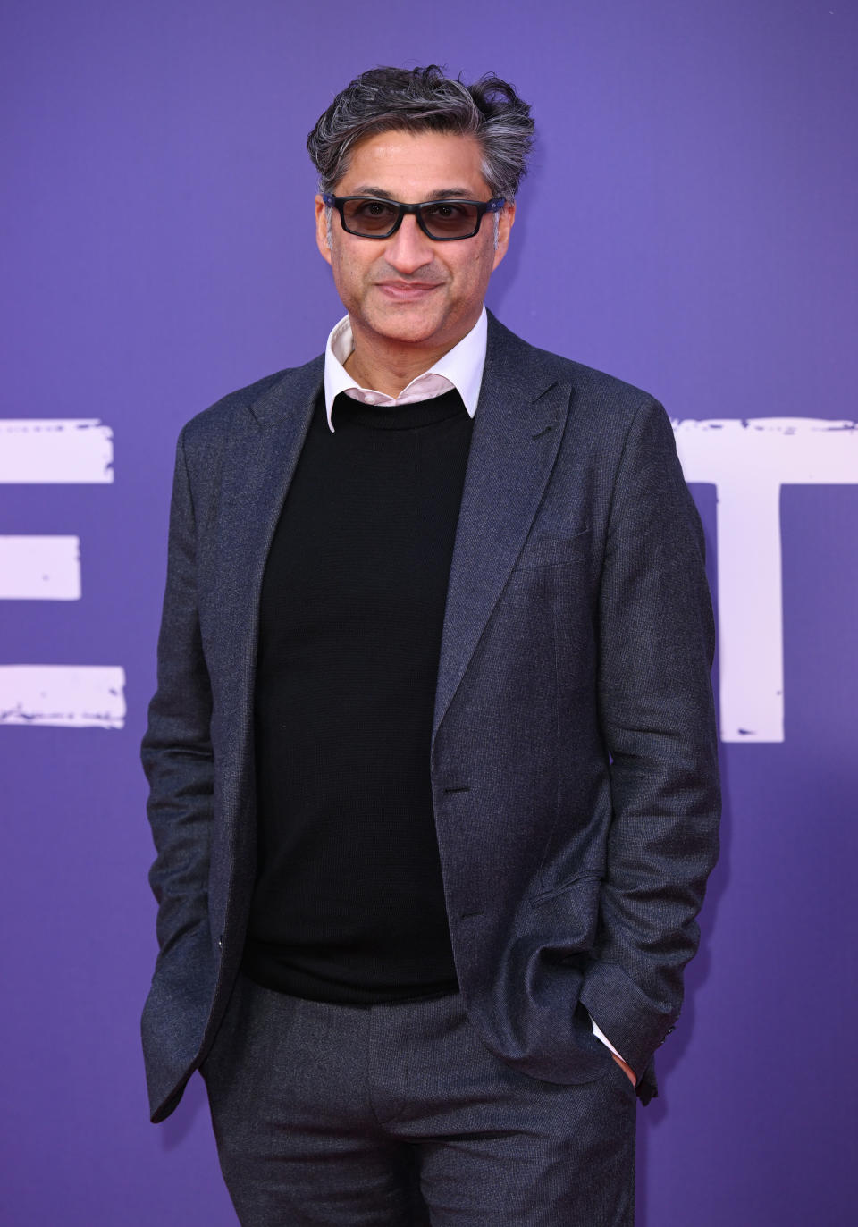 A man in a dark suit, black sweater, and white shirt with his hands in his pockets stands against a backdrop