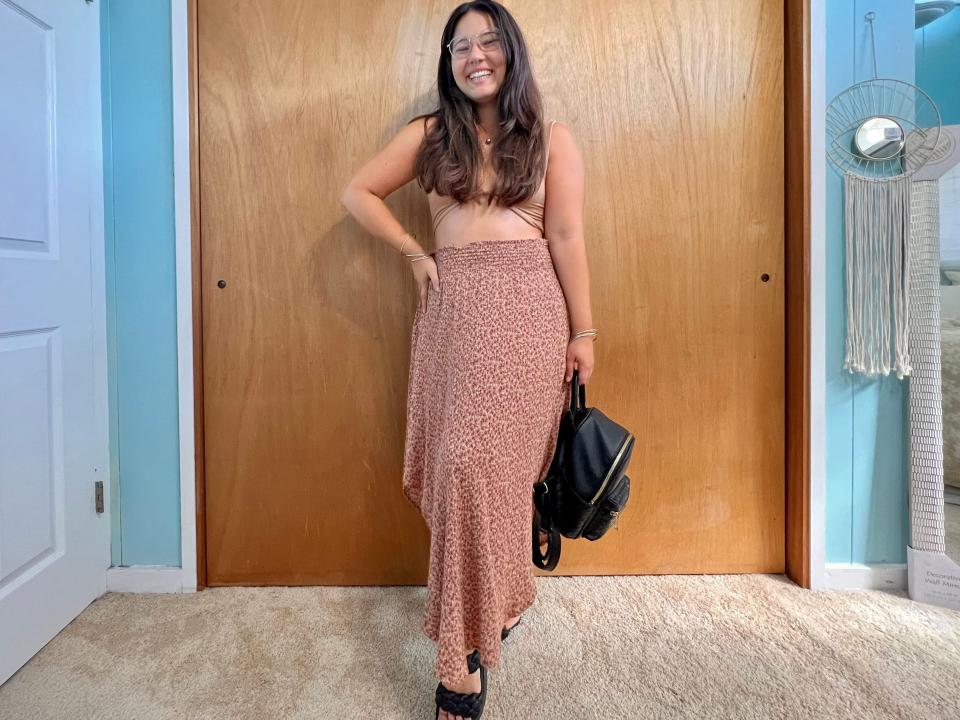 Author poses while wearing tan crop-top, long brown skirt, black backpack, and black sandals.