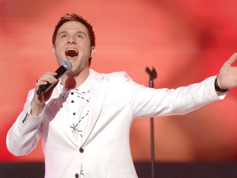 Blake Lewis wears a white suit and holds a microphone and sings in front of a red background