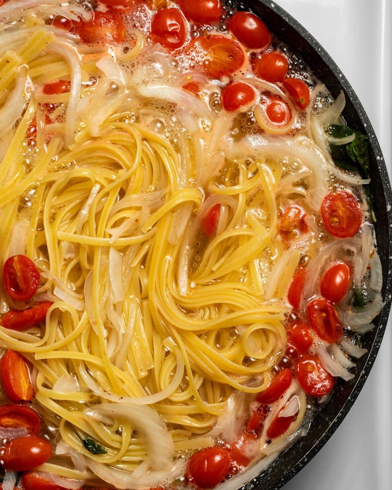 In progress cook with spaghetti noodles, onion, garlic, and cherry tomatoes boiling in water in a cast iron skillet