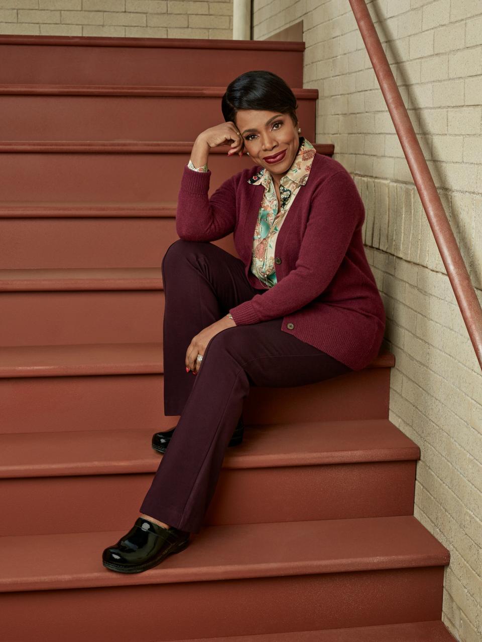 Woman sitting on stairs in a maroon cardigan and pants with a patterned blouse and loafers