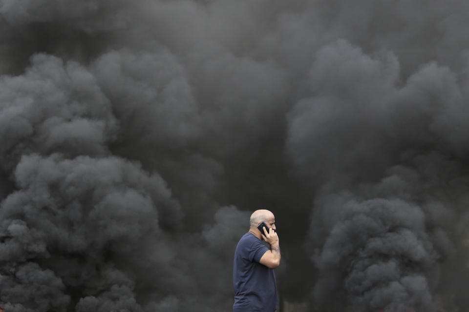 A man speaks on his smartphone while walking by black smoke from burning tires that were set on fire to block a road during a protest against government's plans to impose new taxes in Beirut, Lebanon, Friday, Oct. 18, 2019. The protests erupted over the government's plan to impose new taxes during a severe economic crisis, with people taking their anger out on politicians they accuse of corruption and decades of mismanagement. (AP Photo/Hassan Ammar)