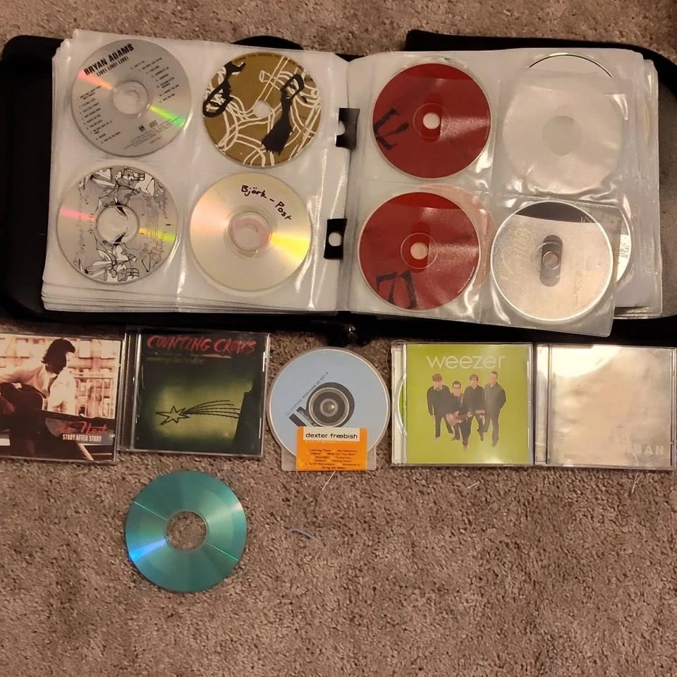 Compilation of various CDs including Weezer in a binder, evoking late '90s to early 2000s music nostalgia