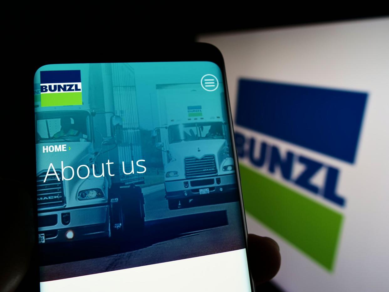 Person holding smartphone with website of British distribution company Bunzl plc on screen in front of logo. Focus on center of phone display.