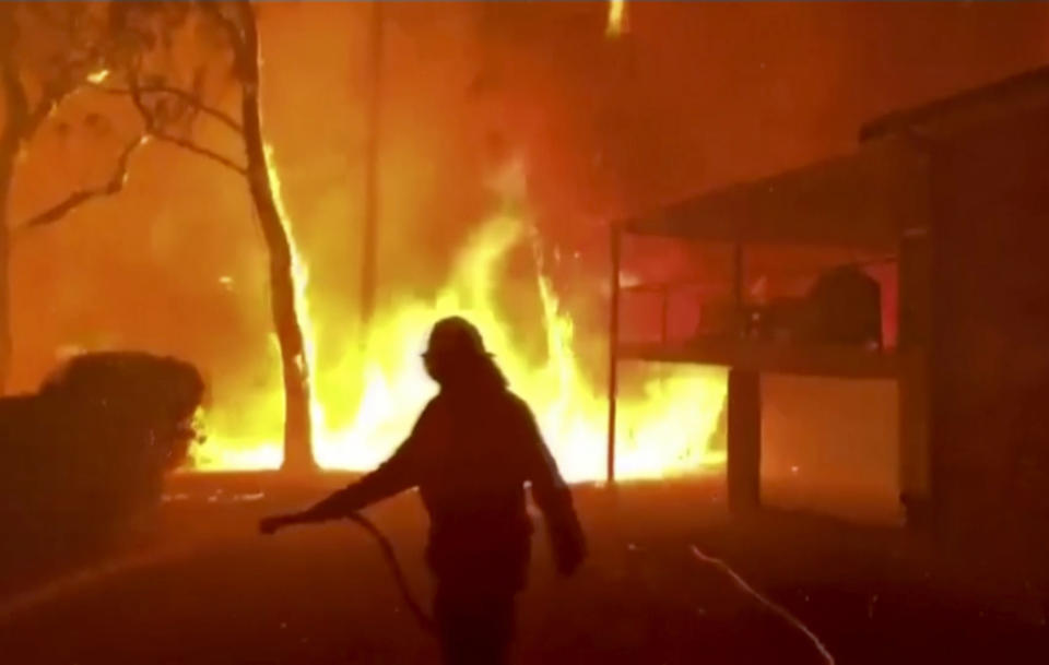 In this image made from video taken and provided by NSW Rural Fire Service via their twitter account, a firefighter sprays water on a fire moving closer to a home in Blackheath, New South Wales state, Australia Sunday, Dec. 22, 2019. Prime Minister Scott Morrison on Sunday apologized for taking a family vacation in Hawaii as deadly bushfires raged across several states, destroying homes and claiming the lives of two volunteer firefighters.(Twitter@NSWRFS via AP)