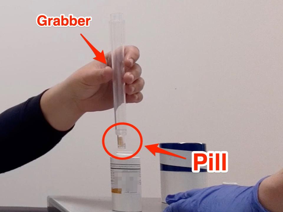 A video still shows Marianne Guenot's hand holding a plastic tube containing the radioactive iodine pill. Arrows point to the grabber and to the pill.