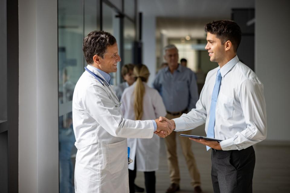 A person in a white lab coat shakes hands with a person holding a tablet.