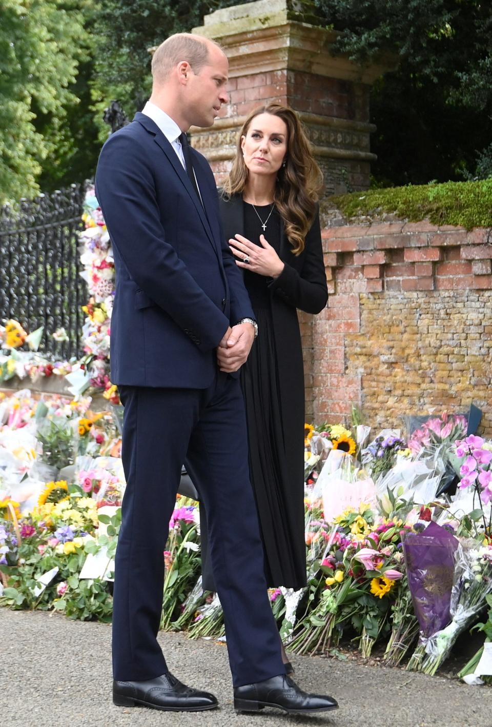 Kate Middleton looks at Prince William with her hand on her heart as they stand in front of flowers.