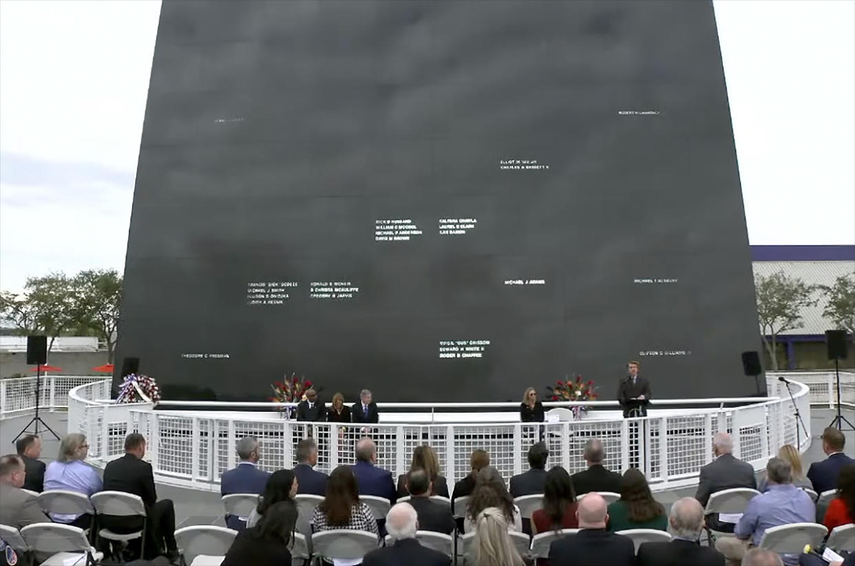  A handful of people in dark suits stand in front of a large black wall into which several dozen names have been carved. 