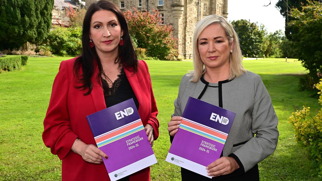 Emma Little-Pengelly and Michelle O'Neill hold copies of the strategy. The backdrop is Stormont Castle, and the couple stands on the lawn. Emma Little-Pengelly wears a red blazer, while Michelle O'Neill wears a black and grey outfit