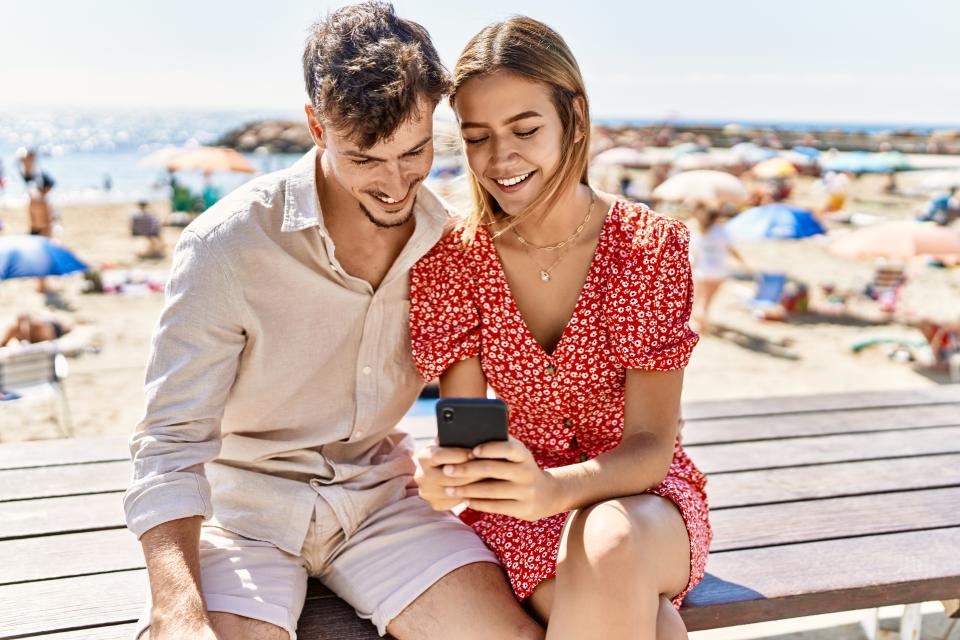 Young hispanic couple on vacation using smartphone sitting on bench at the beach.