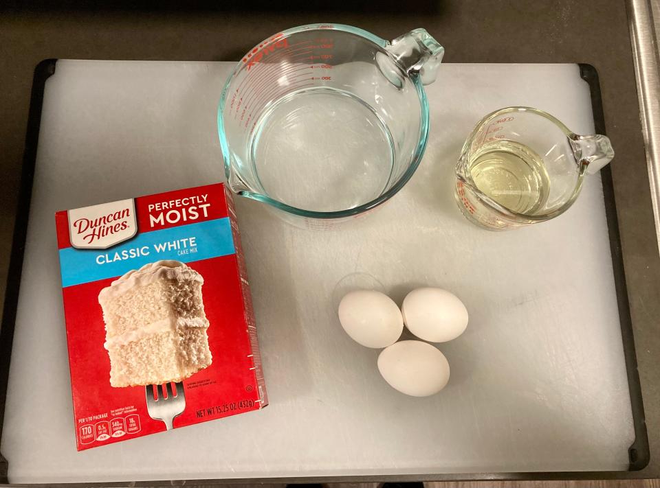 A Duncan Hines vanilla boxed cake mix on a white tray with measuring cups with oil and water and eggs