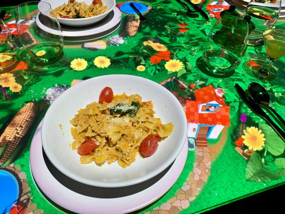 A plate of pasta on a table with projected animations of a garden and a little house.