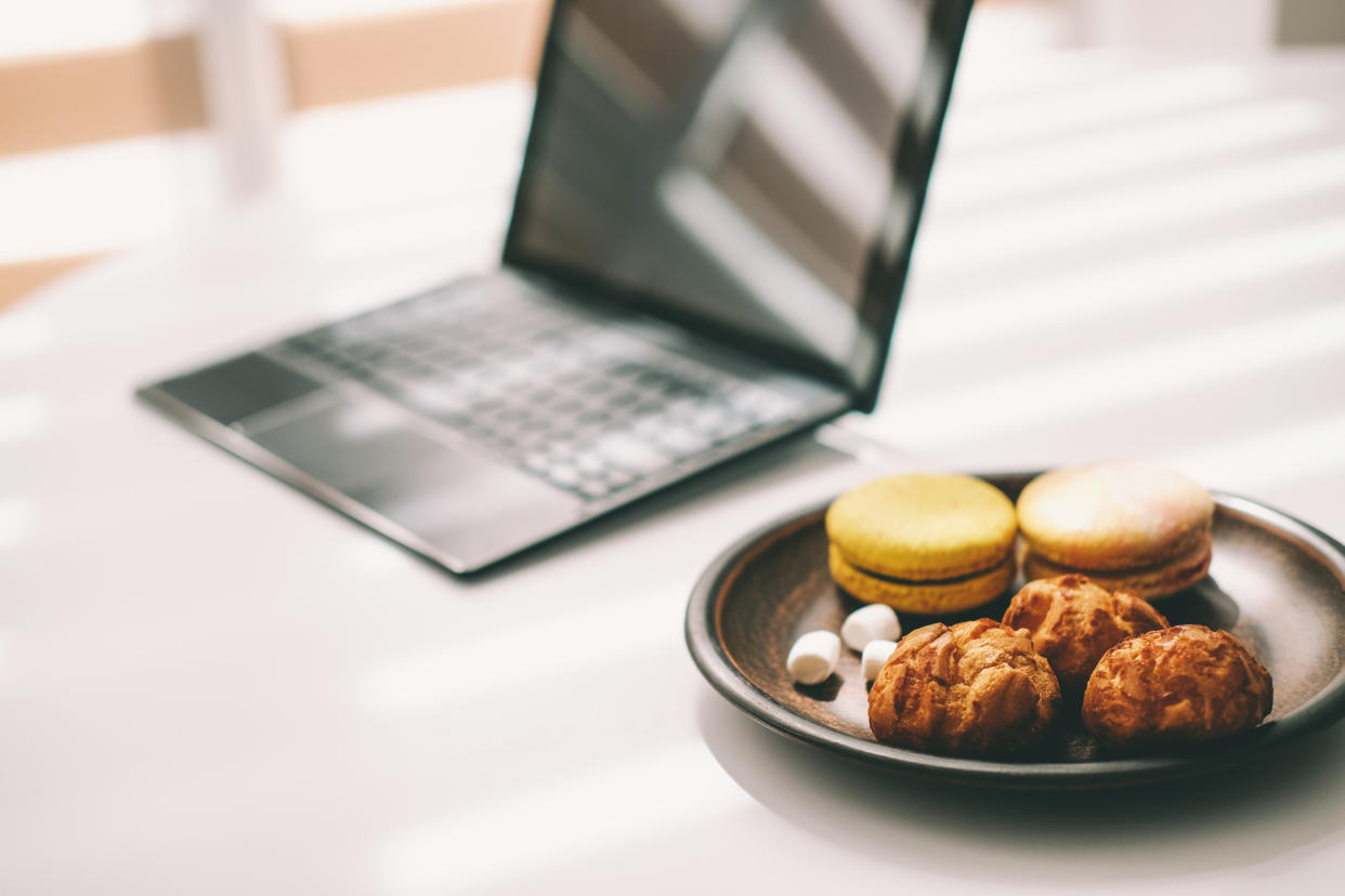 Clay plate of cookies near opened laptop on white table in sunny room with closed blinds.