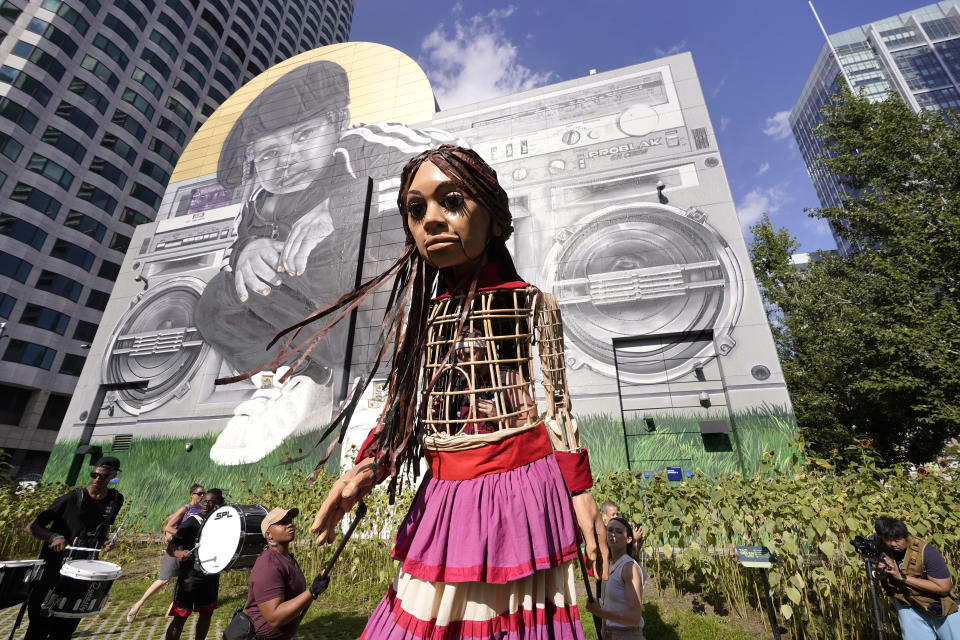 Little Amal, a 12-foot puppet of a 10-year-old Syrian refugee girl, stands in front of a mural with the assistance of puppeteers, Thursday, Sept. 7, 2023, in Boston. The puppet is scheduled to journey across the United States, with planned stops in over 35 towns and cities, between Sept. 7 and Nov. 5, 2023, in an effort to raise awareness about refugees and displaced people across the world. (AP Photo/Steven Senne)