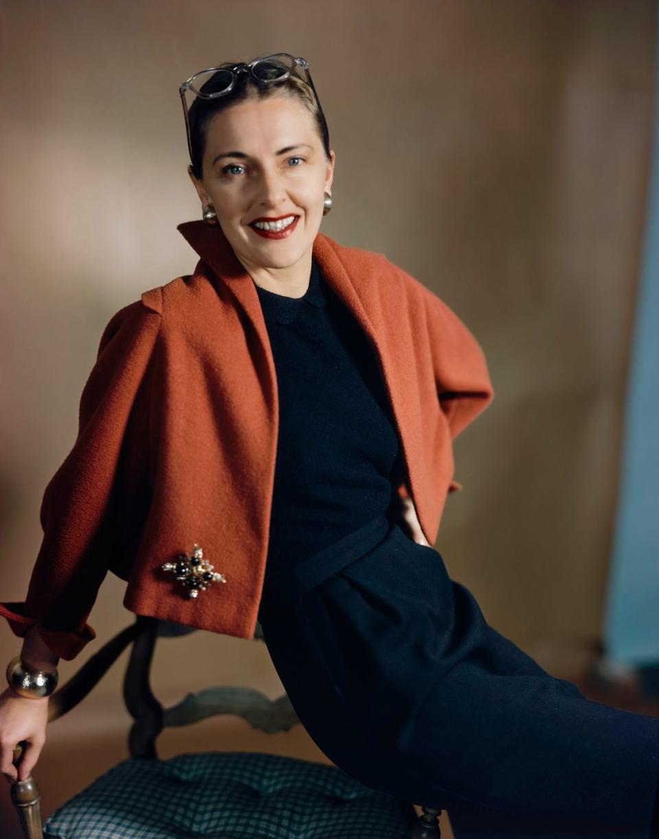 portrait of mrs howard hawks, aka slim keith she is wearing a red bolero capelet by trigere, over a black sheath dress, gold earrings and ring, and a brooch at the hip on the jacket she is seated on the arm of a chair and looking toward the camera, smiling nancy slim hawks vogue february 1, 1949 portrait